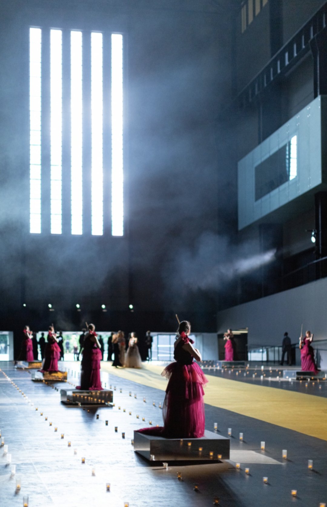 An orchestra in crimson gowns performing among candlelight in a vast darkened hall, staged in the round for an Arcana House production